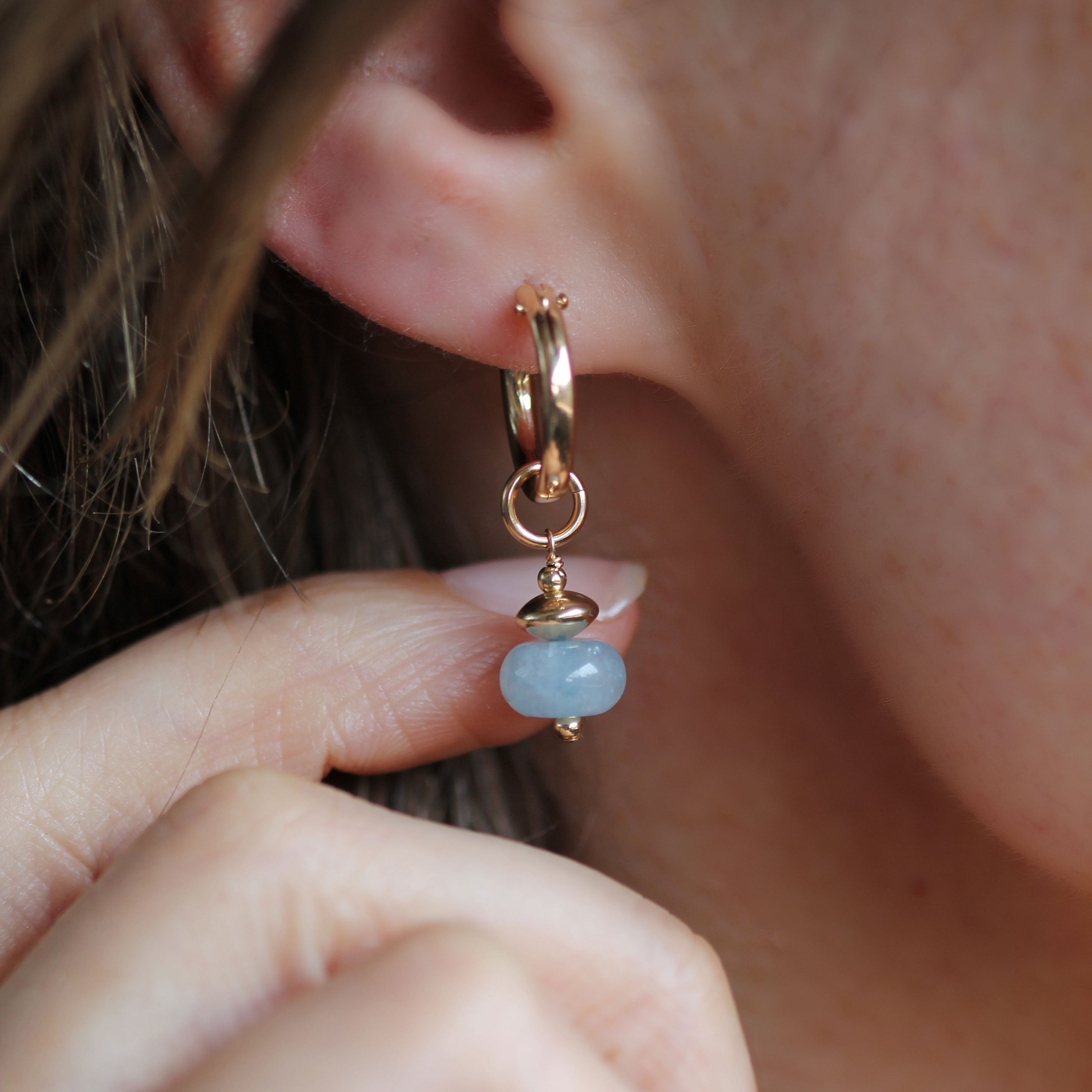 A close-up lifestyle shot of a person wearing a gold huggie hoop earring with a dangling light blue bead charm.