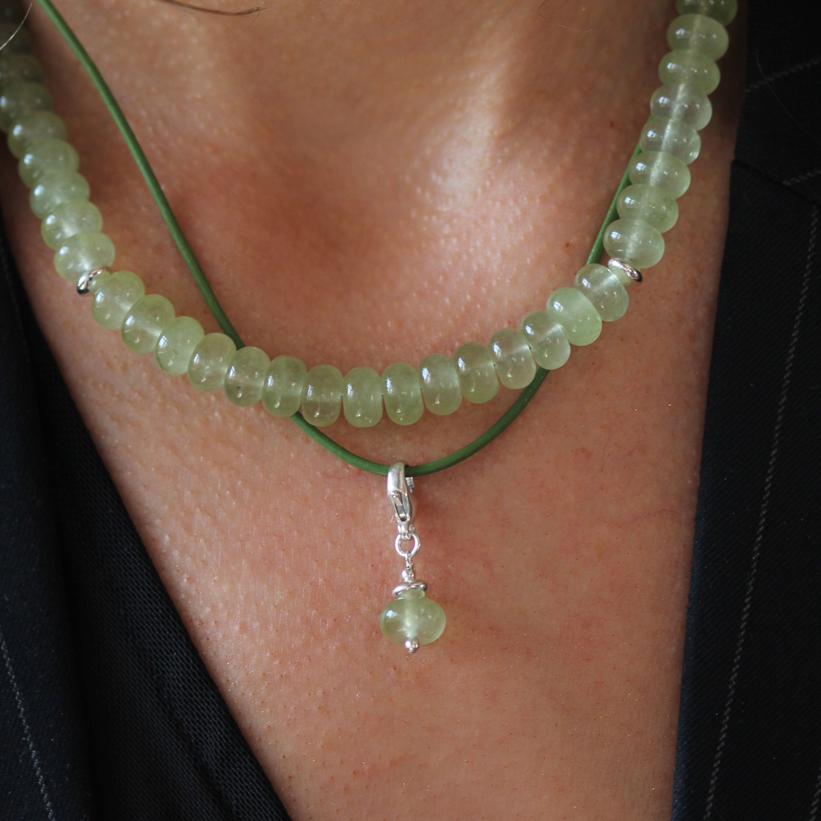 A close-up of a person wearing a double-strand necklace featuring light green beads on one chain and a thin green cord with a small silver pendant containing a green bead on the other.