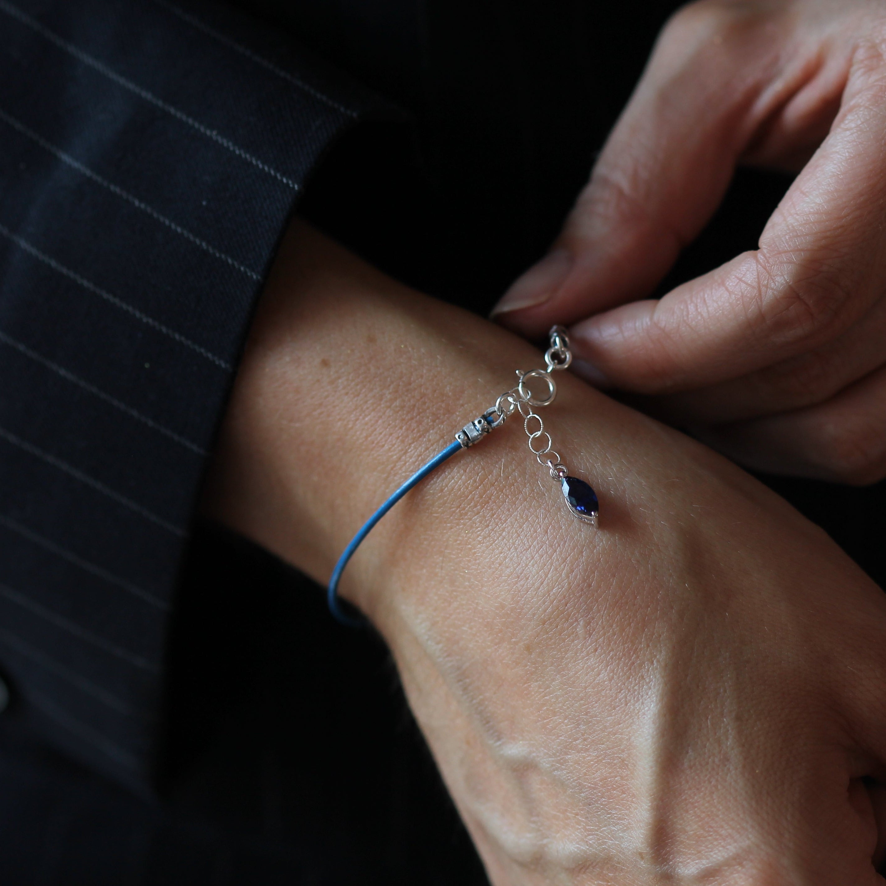 A close-up of a person's wrist wearing a delicate blue cord bracelet with a small silver chain, clear crystal, and dark blue teardrop bead, while another hand adjusts the clasp.
