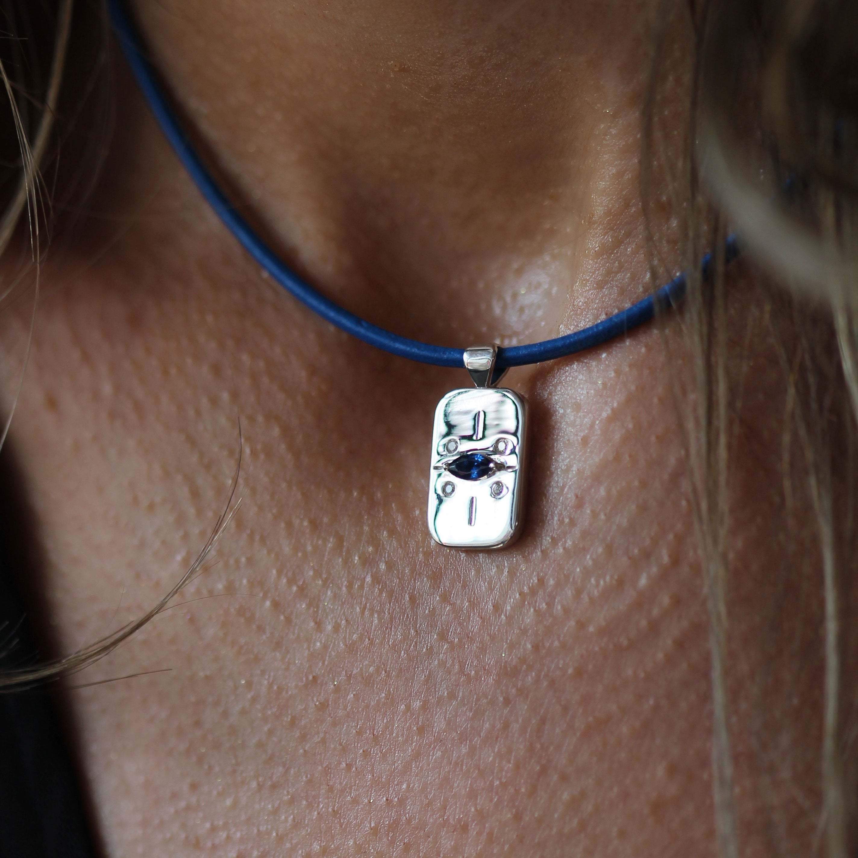 A close-up of a person's neck and chest wearing a silver pendant necklace with a blue cord. The pendant features a skull design with small black and blue details.