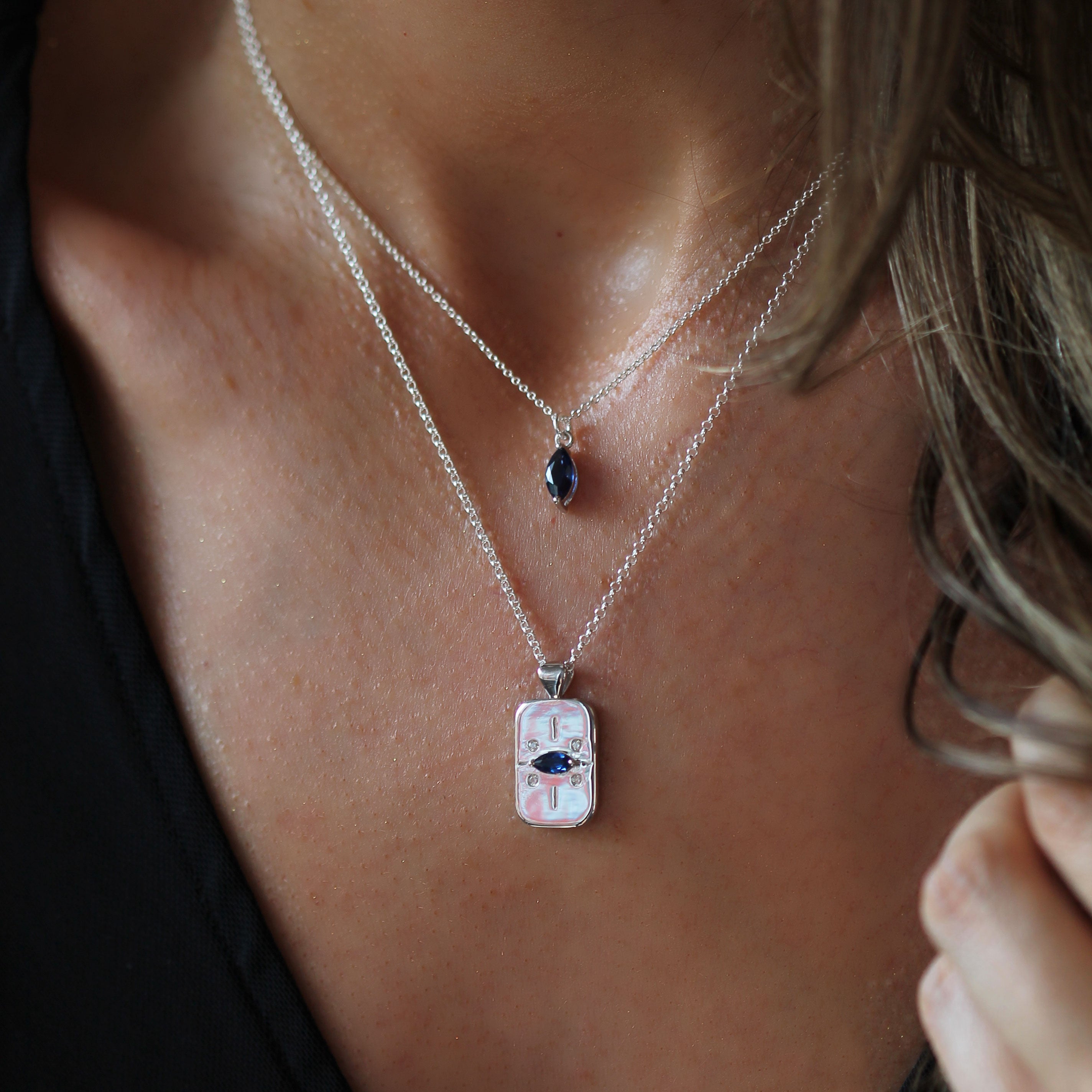 A close-up of a person wearing two layered necklaces. The top necklace has a small blue oval pendant, and the bottom necklace has a silver-toned rectangular pendant.