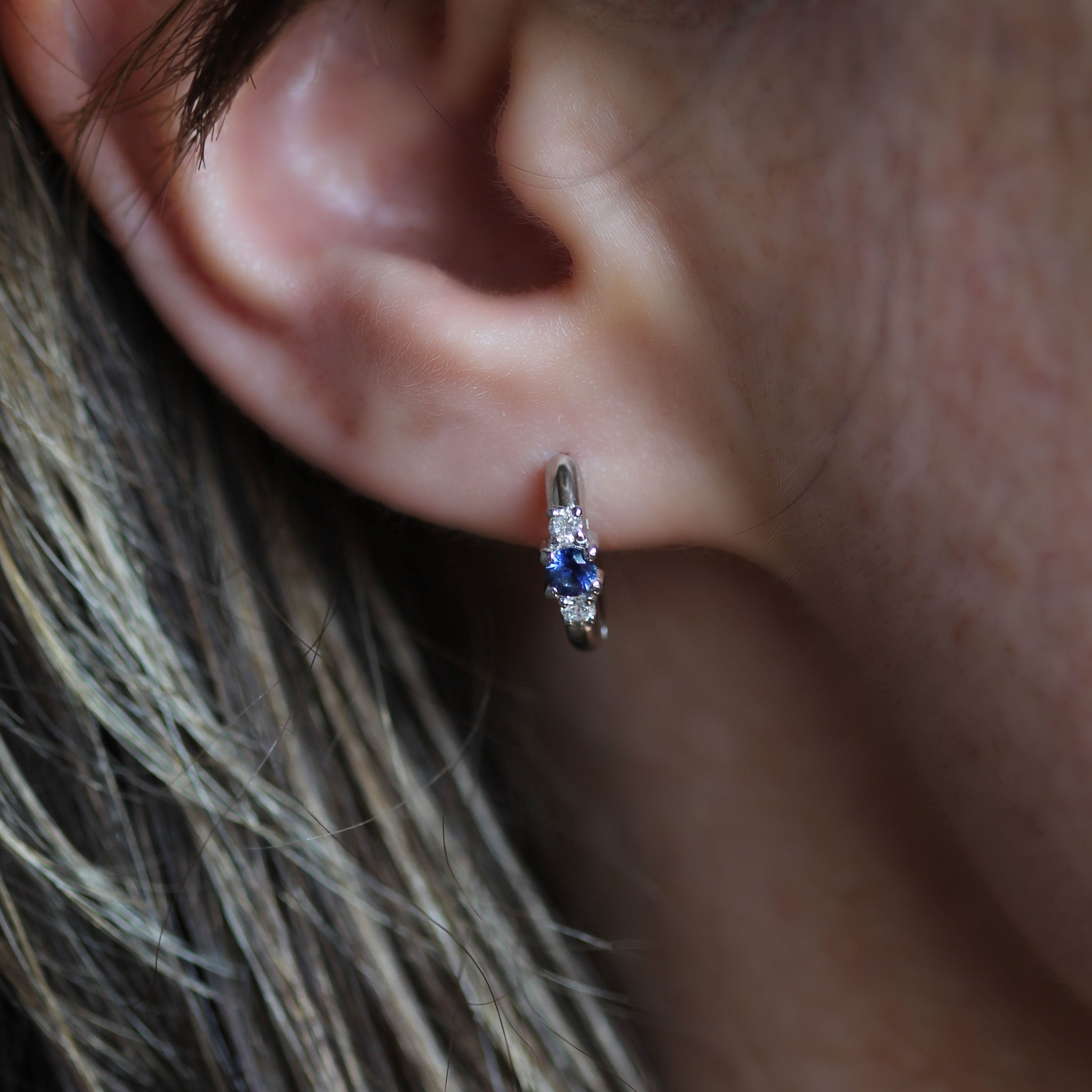 A close-up of a person's ear wearing a small silver huggie hoop earring with a central blue gemstone.