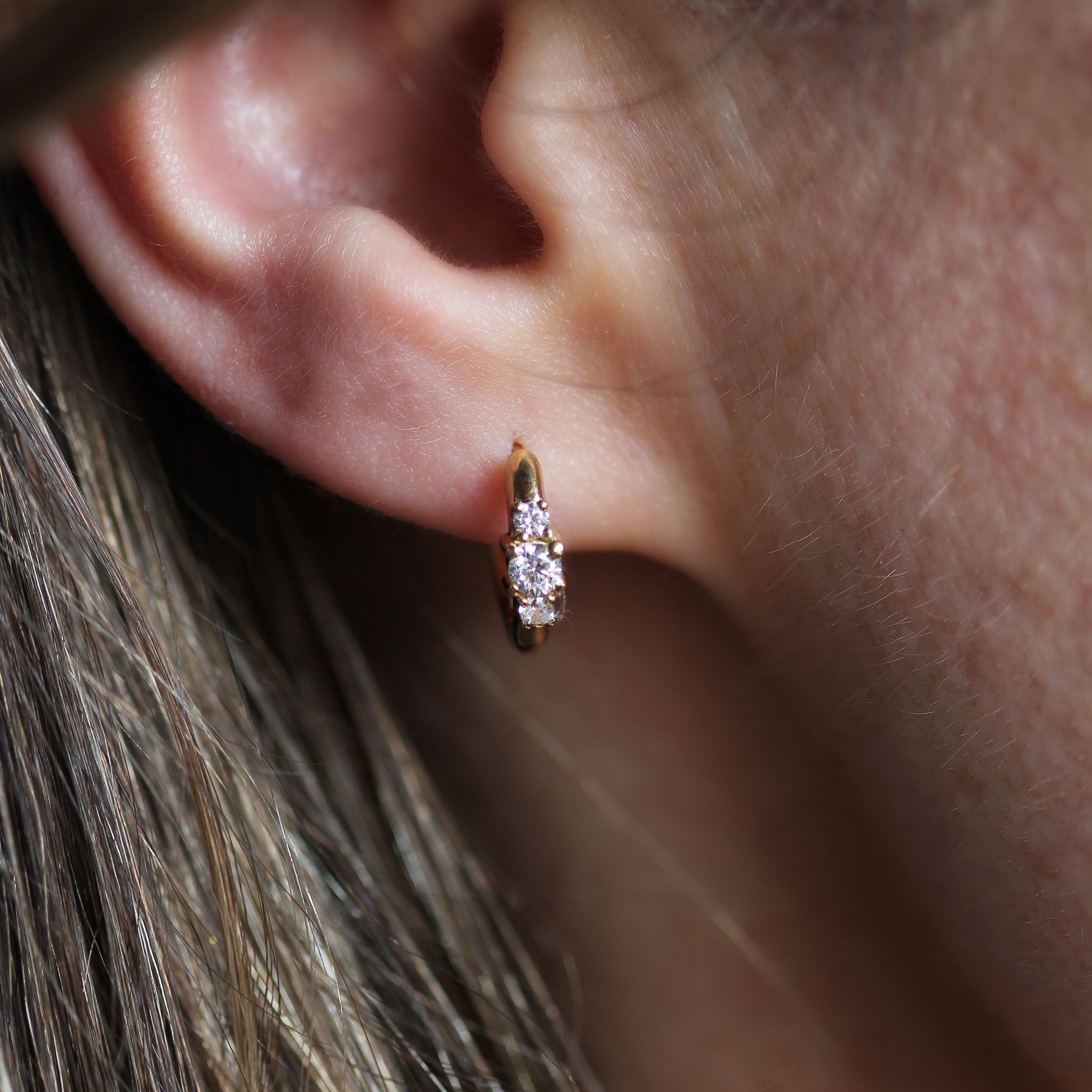A close-up of a person's ear wearing a small gold huggie hoop earring with a clear gemstone.