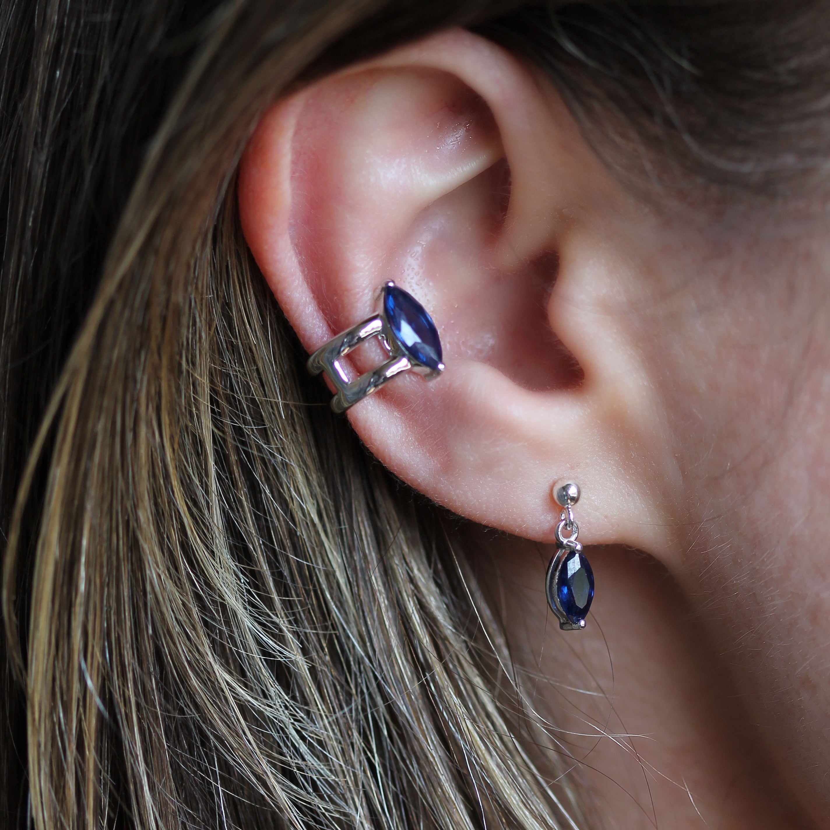 A close-up of a person's ear wearing a silver-toned double-hoop ear cuff with a marquise-cut dark blue gemstone, along with a matching dangling stud earring.