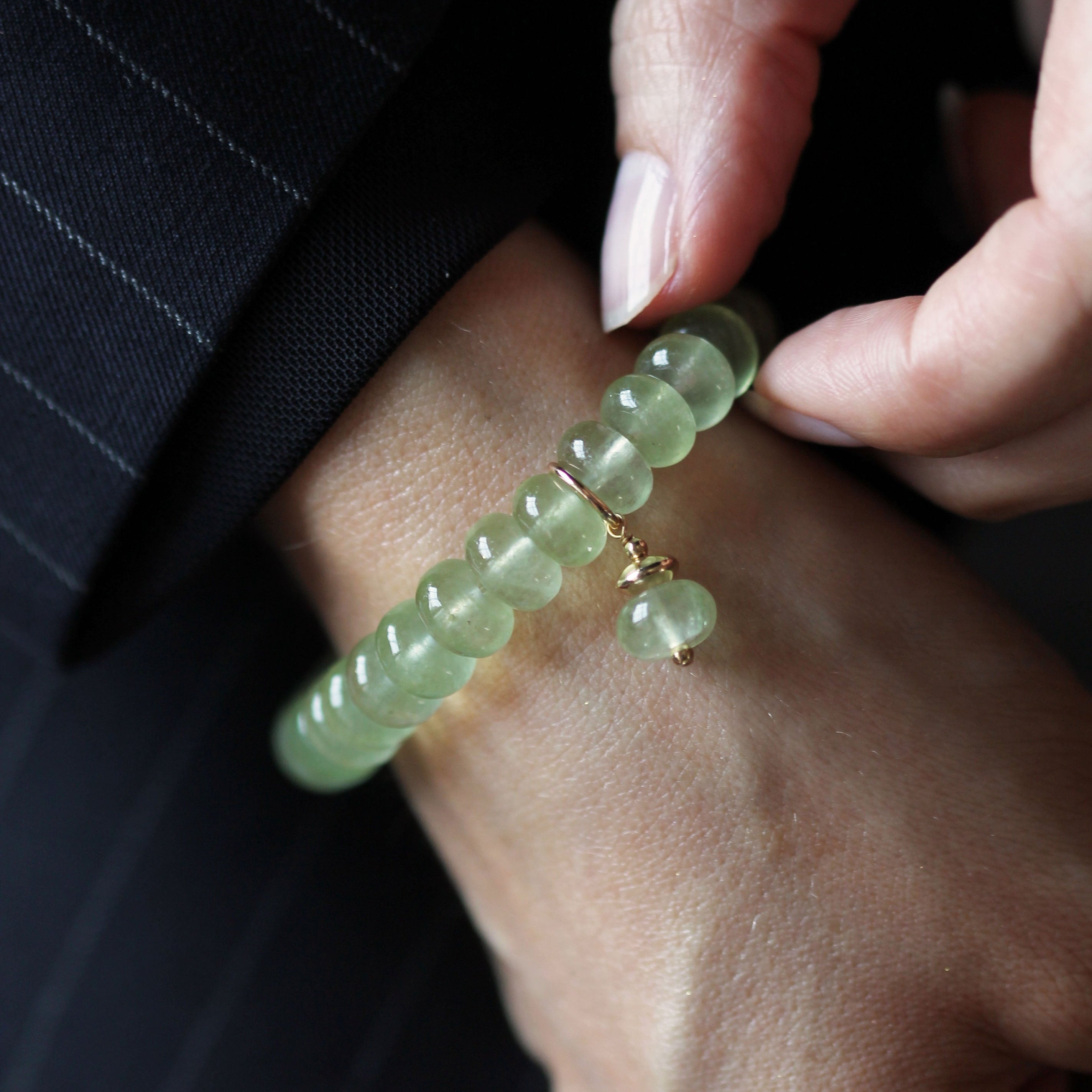 A close-up of a person's wrist wearing a light green beaded bracelet with a small gold charm, while another hand adjusts it. The person is wearing a dark pinstriped jacket.