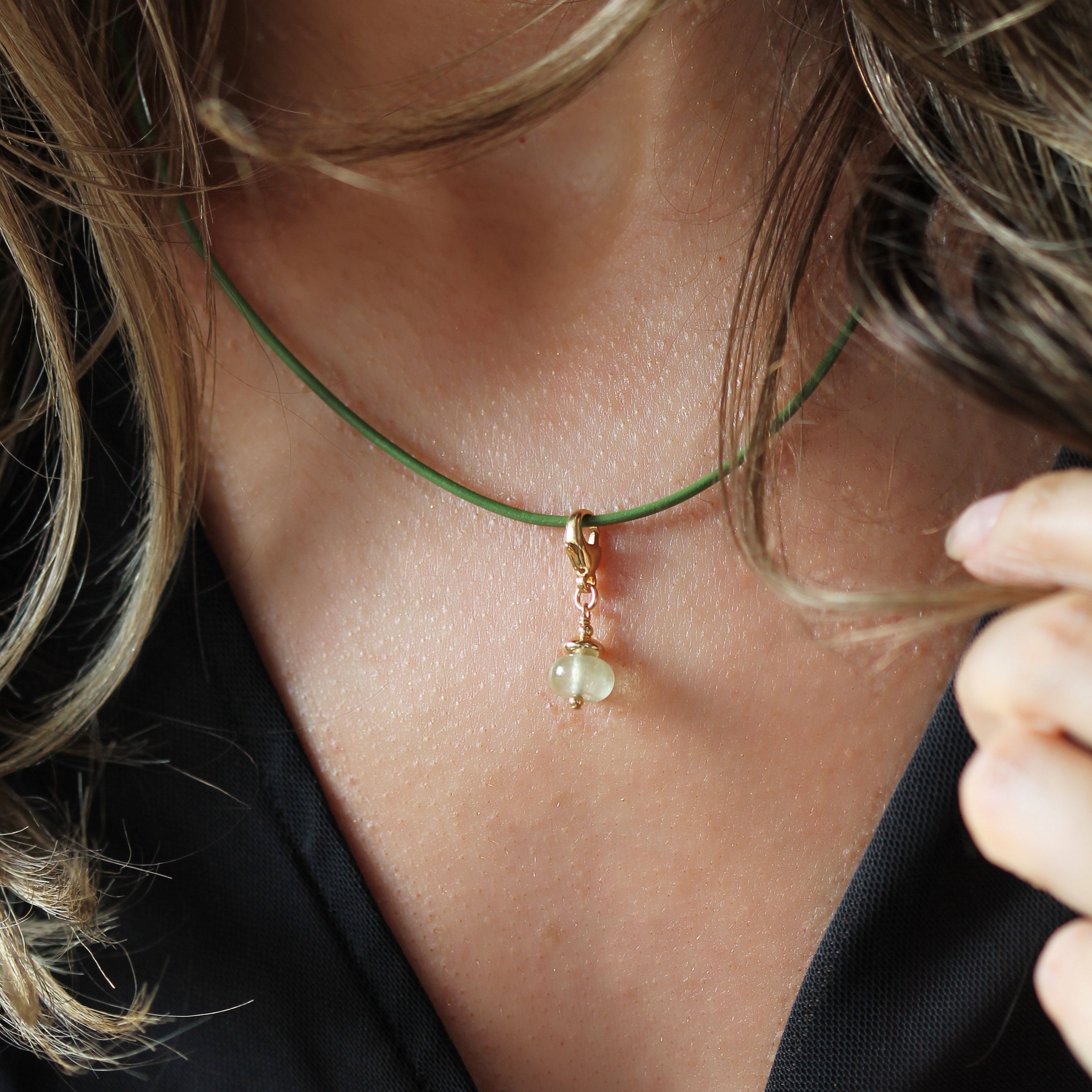 A close-up lifestyle shot of a delicate necklace worn by a model. The necklace features a thin green cord, a small gold bead, and a pale green stone pendant.