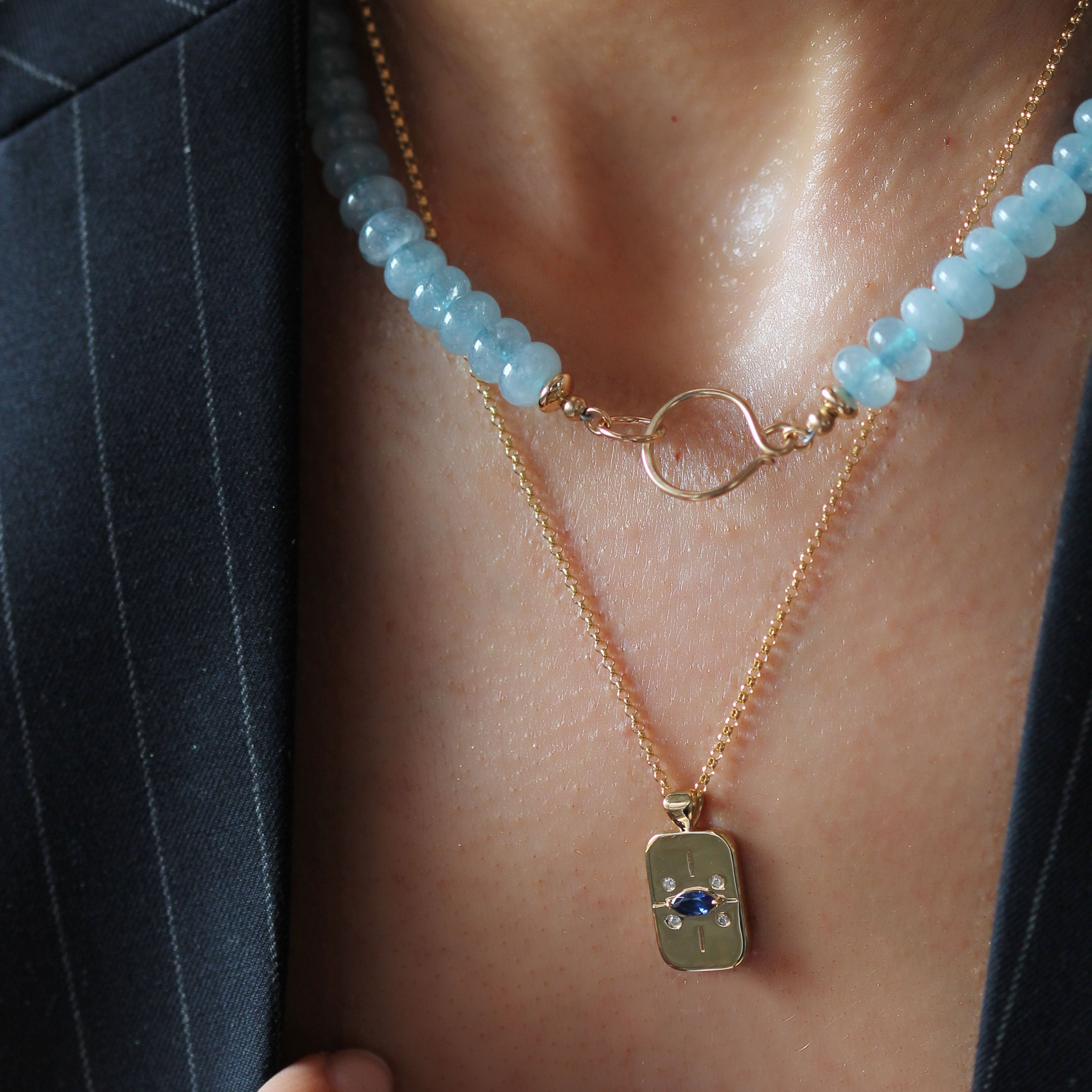 A close-up of a person wearing layered necklaces: a gold chain with a rectangular pendant featuring an eye design, and a second necklace with light blue faceted beads and a gold ring clasp. The person is wearing a dark pinstripe blazer.