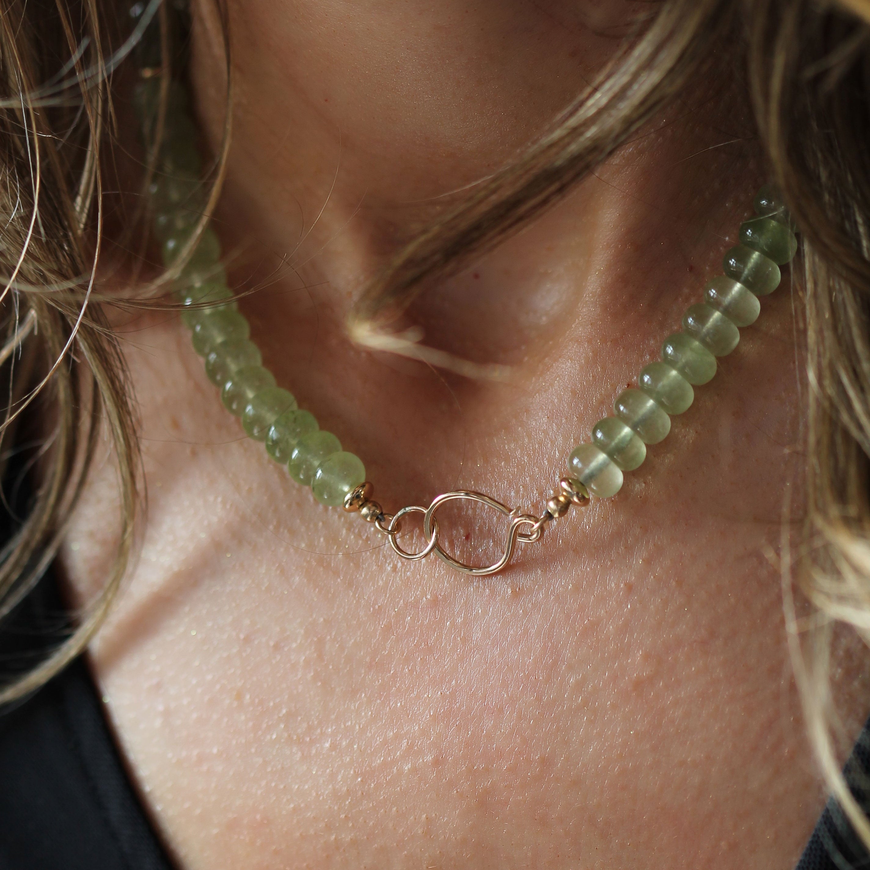 A close-up of a person wearing a beaded necklace with light green faceted beads and a central gold infinity symbol charm.