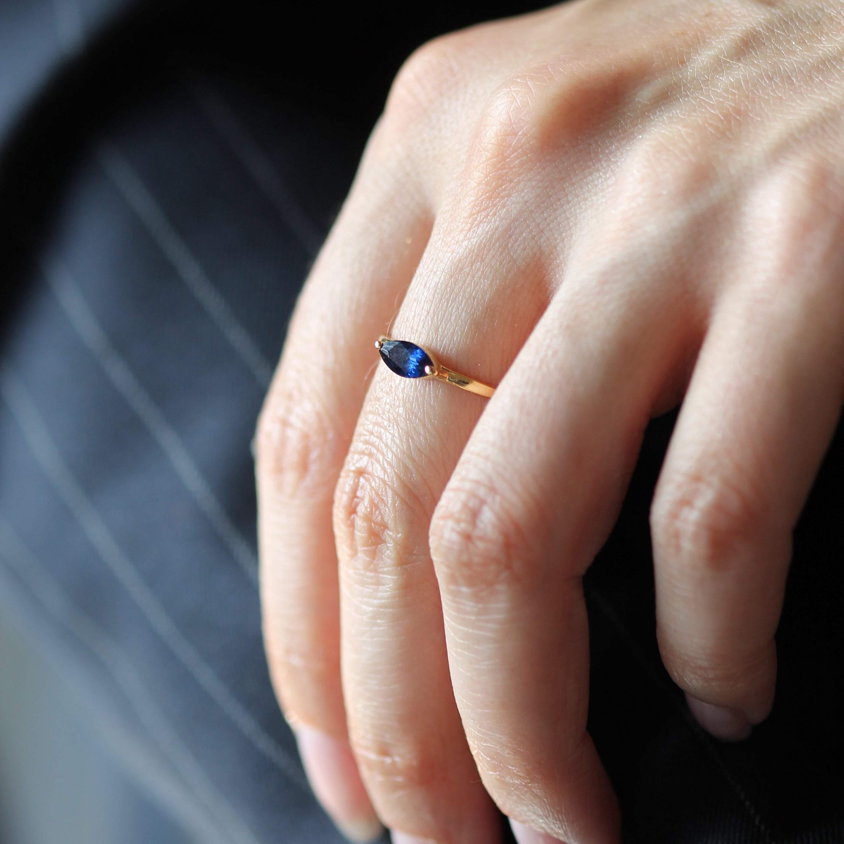 A close-up of a person's hand wearing a gold ring with a marquise-cut dark blue gemstone, resting on a dark pinstriped surface.